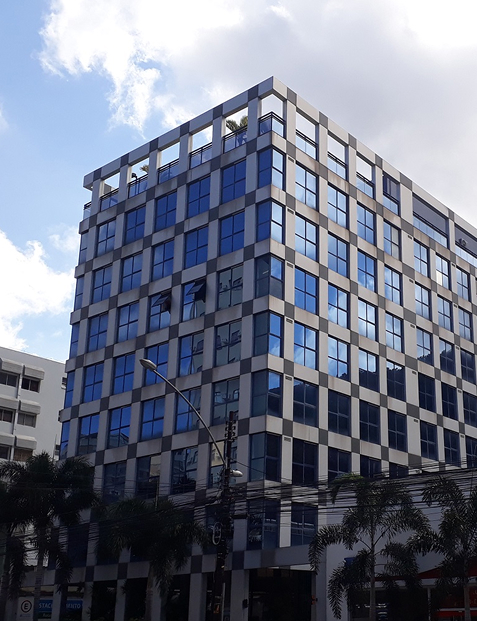 Prédio comercial moderno de fachada envidraçada com estrutura quadriculada, localizado em uma esquina urbana com céu parcialmente nublado ao fundo